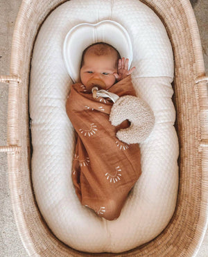 Baby laying down in a baby lounger wrapped in a brixton Phoenix baby swaddle that is the color burnt orange with white sun patterns. Baby has a pacifier and a moon plushy.