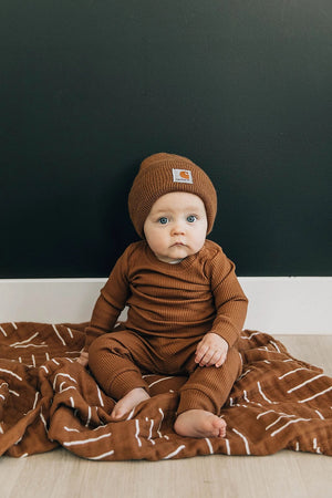 Baby sitting on top of baby swaddle colored rust mudcloth muslin swaddle blanket from mebie baby baby is wearing a rust colored rib two pieces for also wearing a carhart beanie  